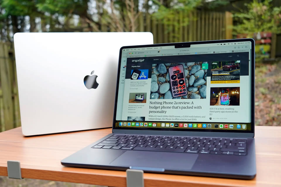 the 2024 macbook air m3 sits on a wooden table outside bear a fence and some trees.
