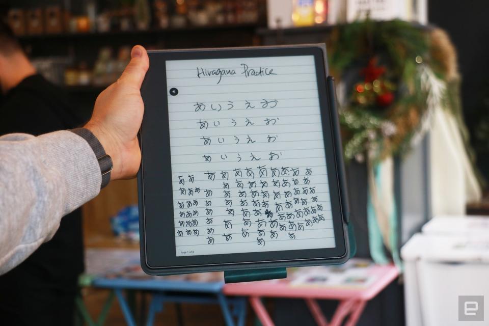 The Amazon Kindle Scribe held up in mid-air by a hand gripping its right side. The screen shows the words