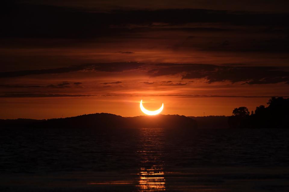 Between layers of clouds, a partial solar eclipse is seen at sunrise over Rice Lake, Ontario, as the sun, partially covered by the moon, rises in a beautiful orange sky above the trees and water.