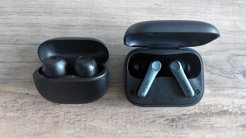 Two black pairs of wireless earbuds, the JLab Go Pop ANC and EarFun Air Pro 4, rest on a light brown wooden table with their cases open, exposing the earbuds as they charge inside.