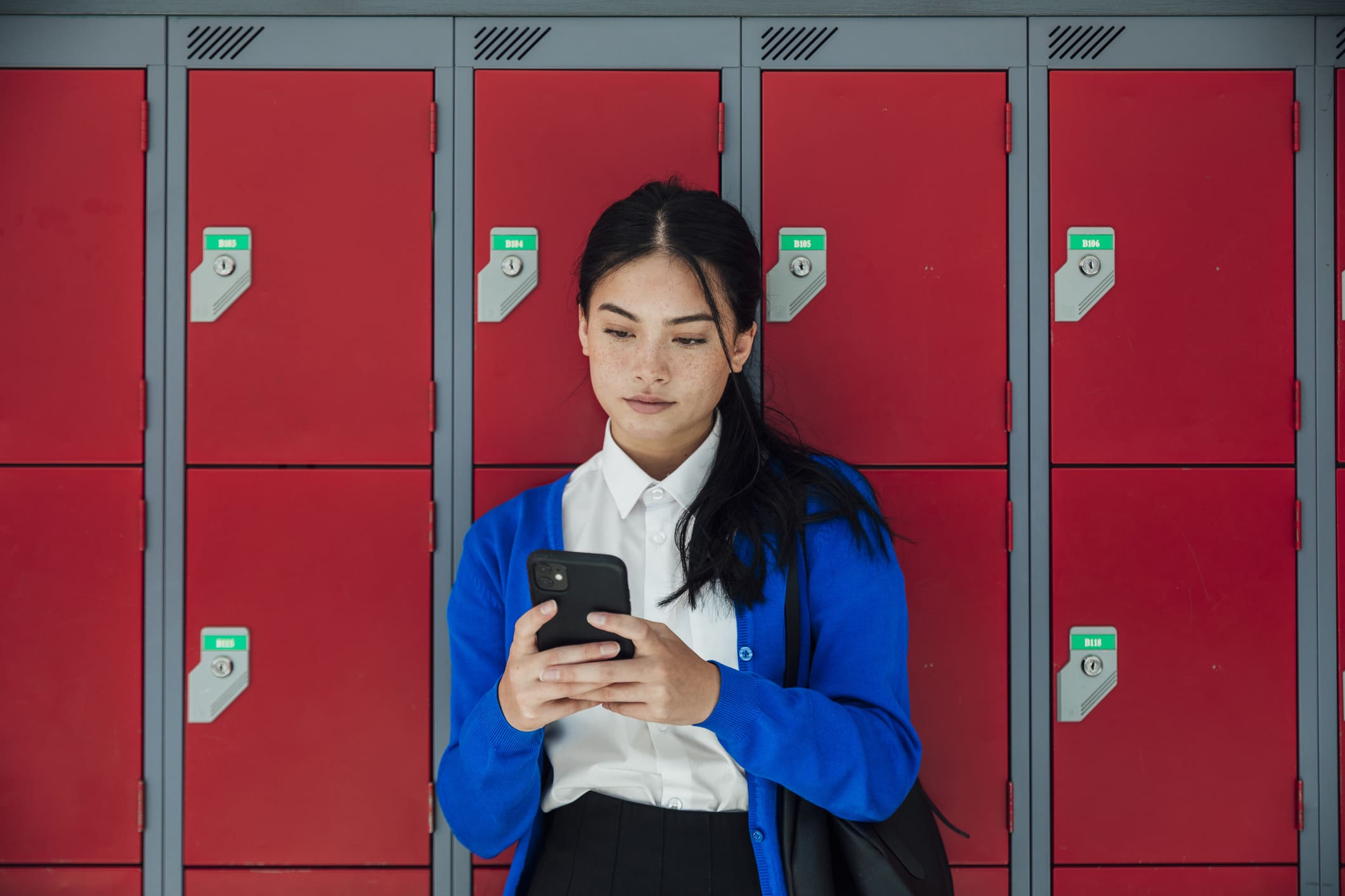 A medium close-up front view of a teenage girl who is sending text messages to a friend and checking her social media as she waits in the corridor of the high school she attends.&nbsp;